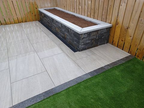 Stone-clad raised planter running along porcelain paving beside an artificial lawn